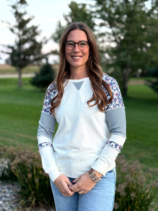 A woman wearing a white long-sleeve top with floral patterned shoulder panels and striped elbow panels, paired with light blue jeans, standing outdoors in a park setting.
