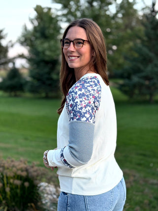 A woman wearing a white long-sleeve top with floral patterned shoulder panels and striped elbow panels, paired with light blue jeans, standing outdoors in a park setting.