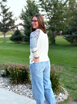 A woman wearing a cream knit sweater with blue and white floral patterned yoke panels and light blue wide-leg jeans, standing outdoors in a park setting.