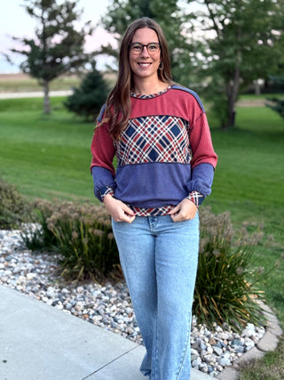 A woman wearing a color-blocked long-sleeve knit sweater with a red and navy blue base and a plaid pattern across the chest, paired with light blue jeans, standing outdoors on a concrete path with a grassy park in the background.