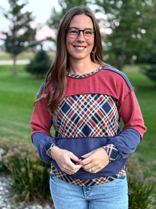 A woman wearing a color-blocked knit sweater with a red, navy, and beige plaid pattern, standing outdoors in a park setting.