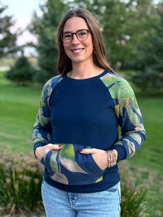 A woman wearing a navy blue crewneck sweatshirt with green and blue abstract leaf patterns on the sleeves and lower body, paired with light blue jeans, standing outdoors in a grassy park setting.