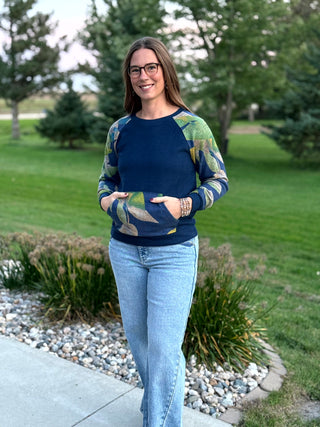 A woman wearing a navy blue long-sleeve raglan sweater with green and yellow abstract leaf patterns on the sleeves stands outdoors on a concrete path in a grassy park setting.