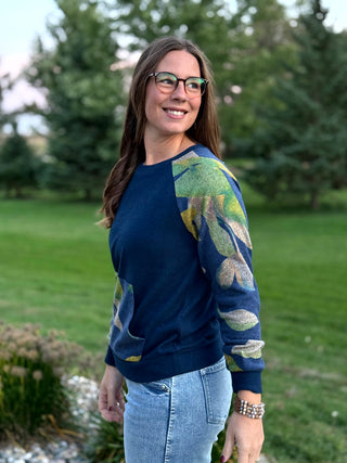 A woman wearing a navy blue crewneck sweatshirt with green and gold glittery leaf-patterned sleeves, paired with light blue jeans, standing outdoors in a grassy park setting.