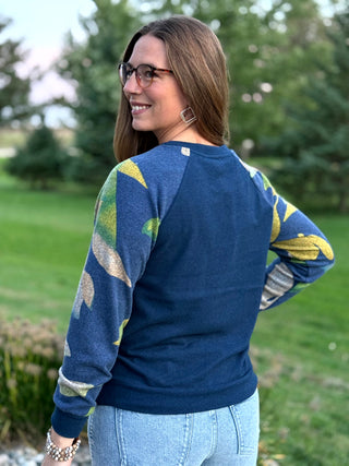 A woman wearing a navy blue long-sleeve raglan sweatshirt with abstract green, yellow, and gray patterned sleeves, photographed outdoors on a grassy lawn.