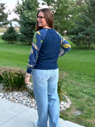 A woman wearing a navy blue raglan sweatshirt with green and yellow patterned sleeves, paired with light blue jeans, standing outdoors on a concrete path with a grassy lawn and trees in the background.