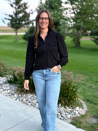 Full front view of a model wearing a black long-sleeve quarter-zip blouse with light-wash jeans.