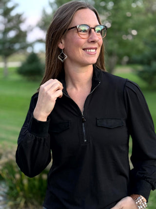 Front detail of a black blouse featuring a quarter-zip neckline, flap chest pockets, and pleated shoulders.