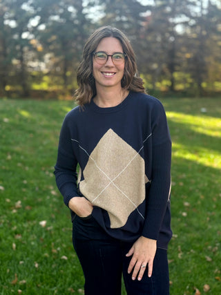 Model wearing a navy sweater with a large beige argyle diamond, cream diagonal lines, and ribbed sleeves.