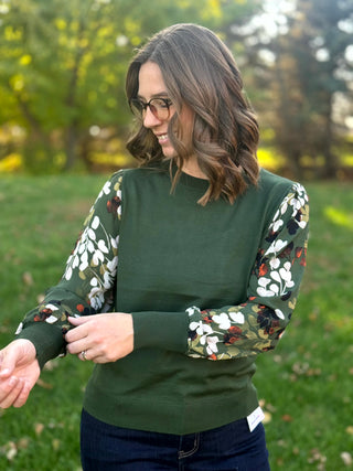 Model adjusting the cuff of a forest green crewneck top with gathered leaf-print sleeves.
