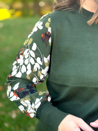 Close-up of a forest green top showing the knit bodice, leaf-print sleeve, and wide ribbed cuff.