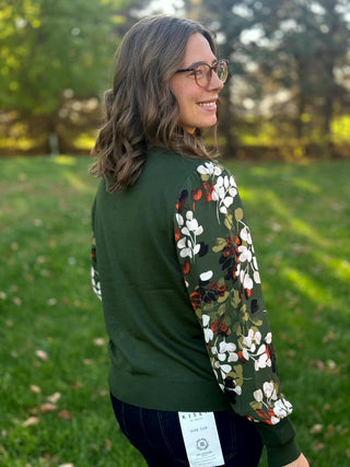 Rear three-quarter view of a forest green top with multicolor leaf-print sleeves and ribbed hem.
