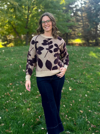Model wearing a taupe sweater with dark plum leaf motifs, cream ribbed trim, and dark wide-leg jeans, front view.