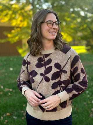 Close-up front view of a taupe knit sweater with dark plum leaves, a crew neckline, and cream ribbed cuffs and hem.