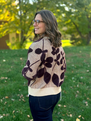 Angled back view of a taupe sweater showing dark plum leaf motifs, dropped shoulders, and a cream ribbed hem.