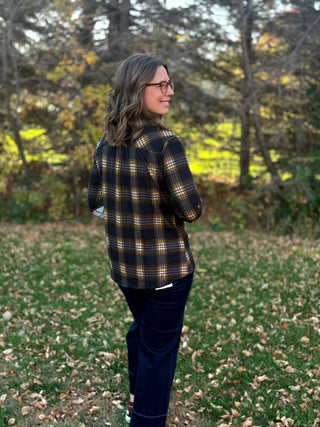 Back view of a model wearing a black, tan, and gray plaid shirt with a curved hem and dark jeans.