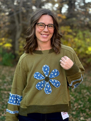 Front view of olive green crewneck sweatshirt with frayed blue floral flower appliqué, patterned sleeve bands, and dark olive ribbed trim.