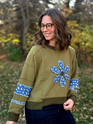 Angled front view of relaxed olive green sweatshirt with blue floral flower appliqué and matching sleeve accents.