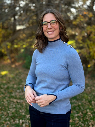 Front view of a light blue mock-neck sweater with gold-tone shoulder buttons and navy trim at the neckline and split cuffs.