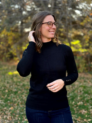 Front view of a black mock-neck sweater with long sleeves, ribbed hem, and decorative cuff buttons.