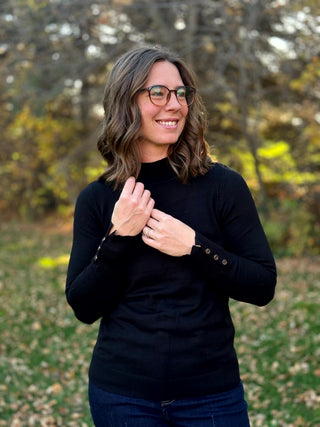Close-up of a black mock-neck sweater showing decorative buttons on both ribbed cuffs.