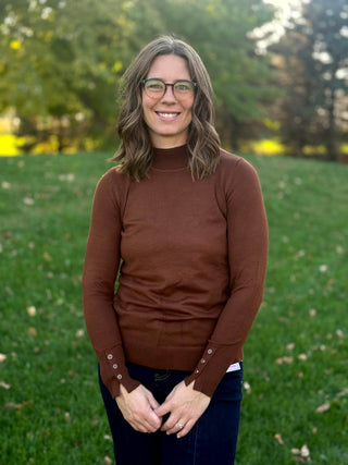 Front close-up of a brown mock-neck sweater showing its fitted silhouette, ribbed hem, and button-detail cuffs.