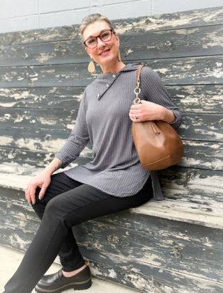 Woman sitting on a wooden bench wearing a gray top and black pants, holding a brown handbag.