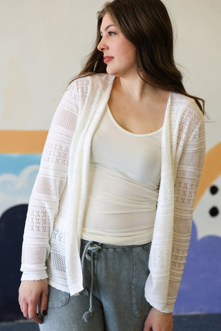 Woman wearing a white lace cardigan over a white top and gray pants, standing indoors.