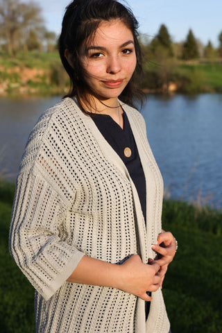 Woman wearing a light gray cardigan standing by a body of water with trees in the background