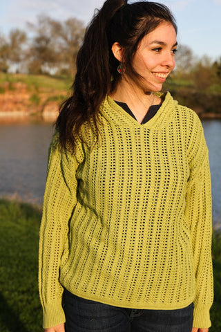 Woman wearing a green sweater standing by a body of water with trees in the background