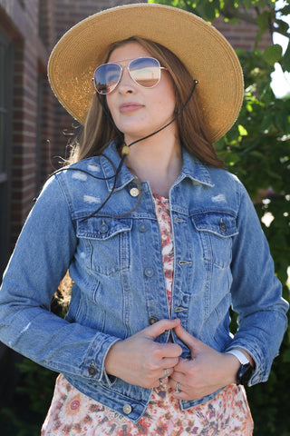 Woman wearing a denim jacket, floral dress, and straw hat outdoors.