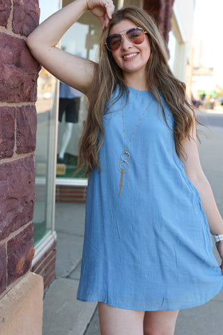 Woman in a blue dress and sunglasses leaning against a brick wall.